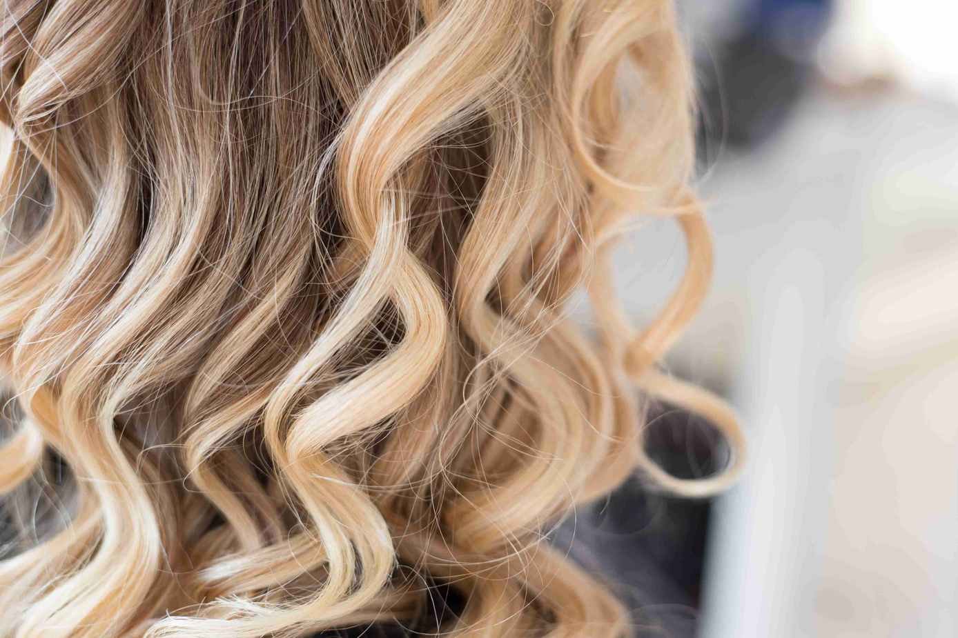 Blond loki wypełniają kadr, miękko skręcone, na rozmytym jasnym tle.