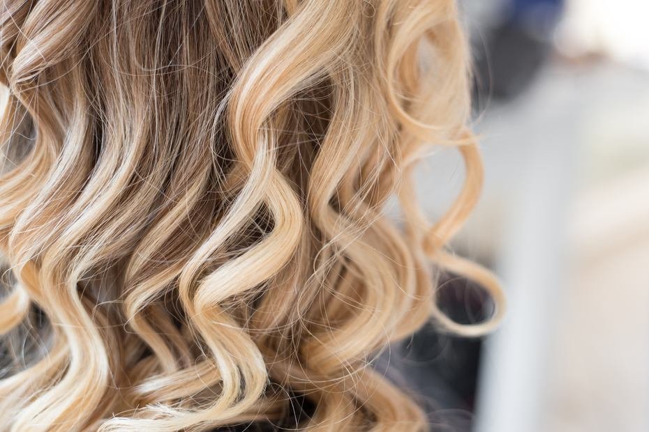 Blond loki wypełniają kadr, miękko skręcone, na rozmytym jasnym tle.