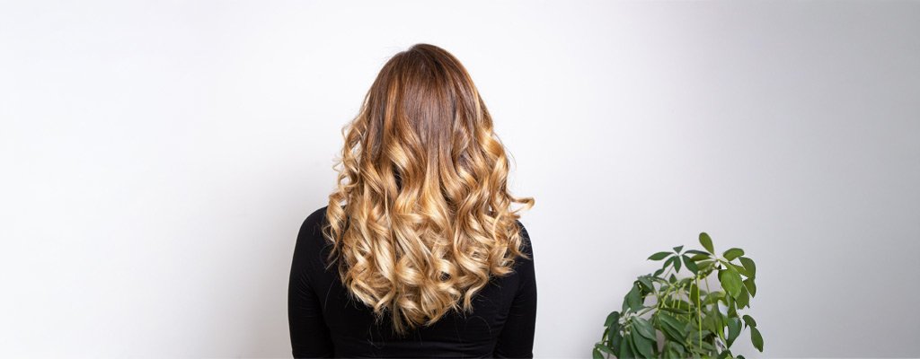 Kobieta od tyłu prezentuje długie, falowane blond włosy na białym tle.