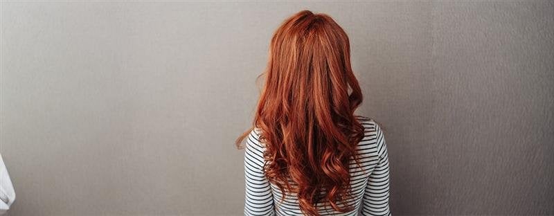 Kobieta z długimi rudymi włosami stoi tyłem przy szarej ścianie.