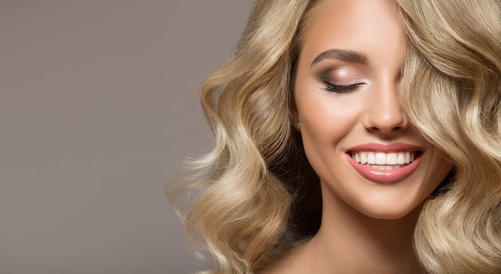 Portret uśmiechniętej blondynki z włosami koloru blond do ramion, na szarym tle.