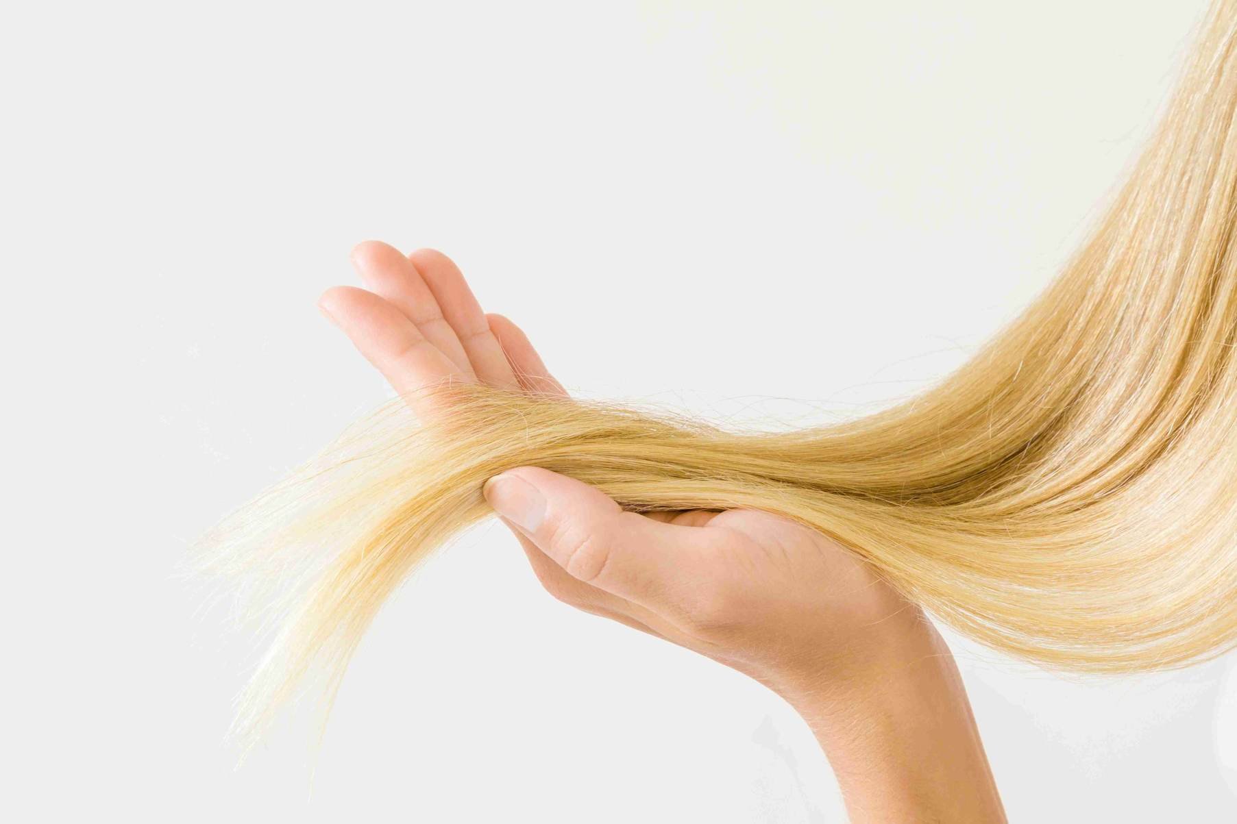 Dłonie trzymają i wygładzają blond pasmo włosów na jasnym tle.
